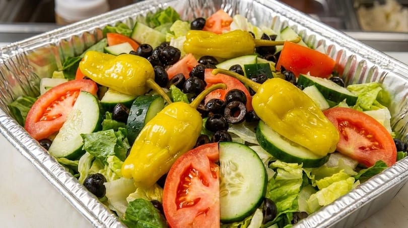 Fresh garden salad in a foil container with lettuce, tomatoes, cucumbers, black olives, and yellow peppers.