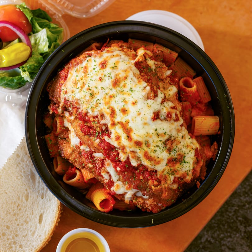 Overhead view of chicken parmesan with rigatoni, salad, bread, and dressing on a wooden table.