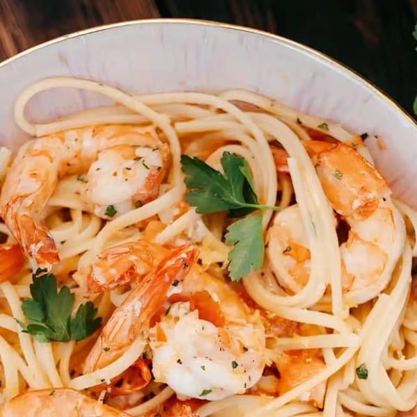 Close-up of shrimp scampi with linguine pasta, tomatoes, and parsley in a white bowl.