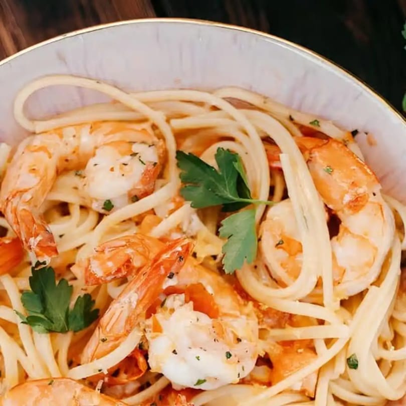 Close-up of shrimp scampi with linguine pasta, tomatoes, and parsley in a white bowl.